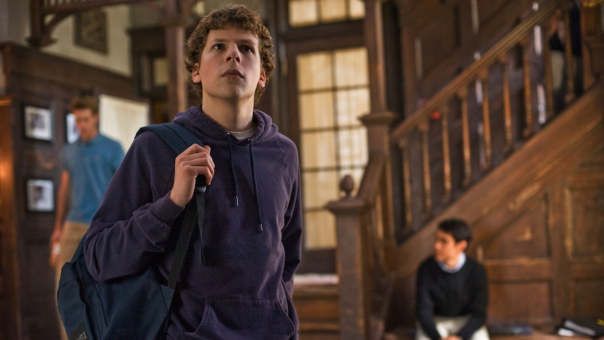 Jesse Eisenberg stands in a foyer wearing a blue hoodie and a black backpack slung over one shoulder.