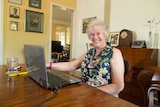 An older woman sits in front of a laptop at her dining room table.