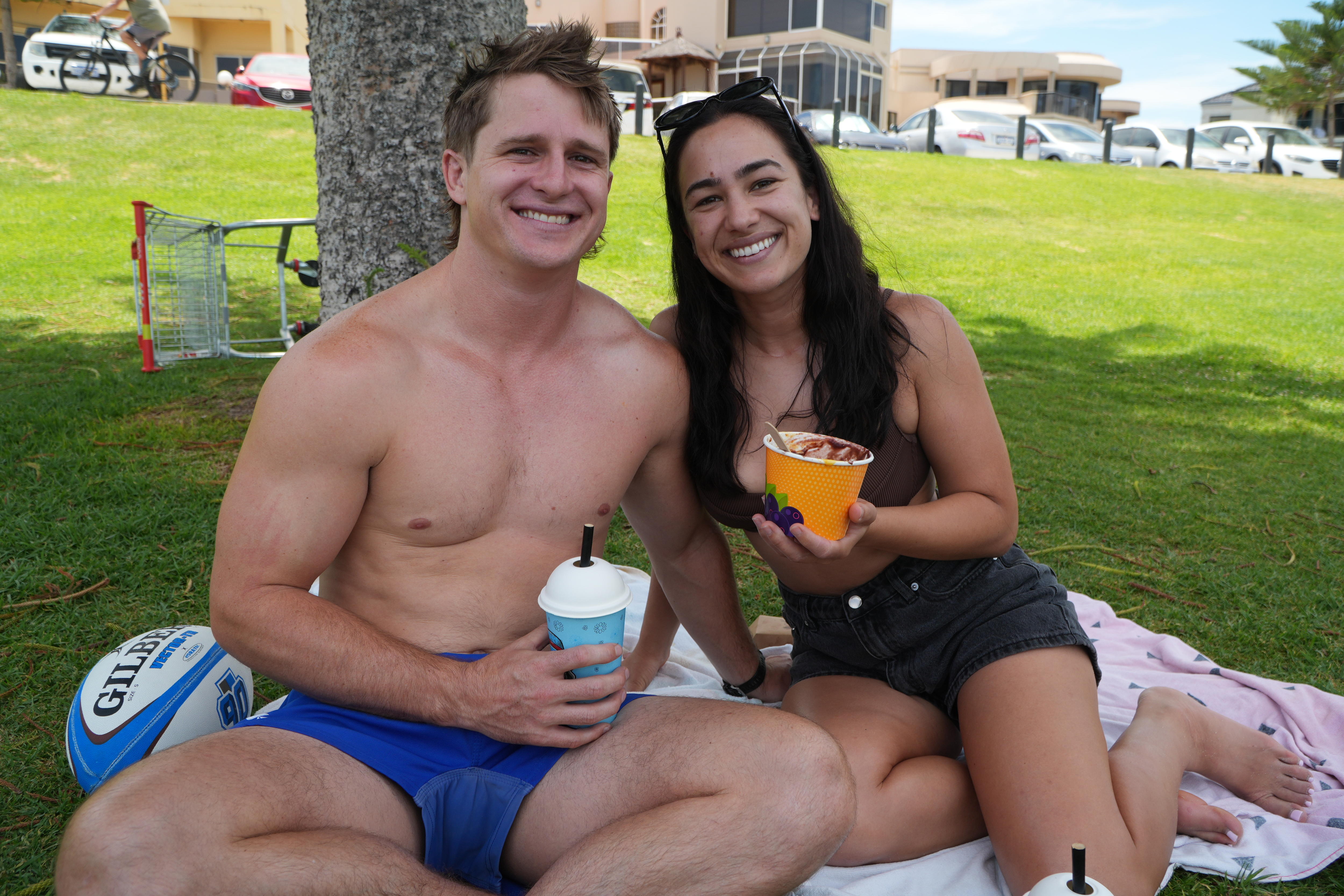 A youthful couple is seated on a grassy patch beaming.
