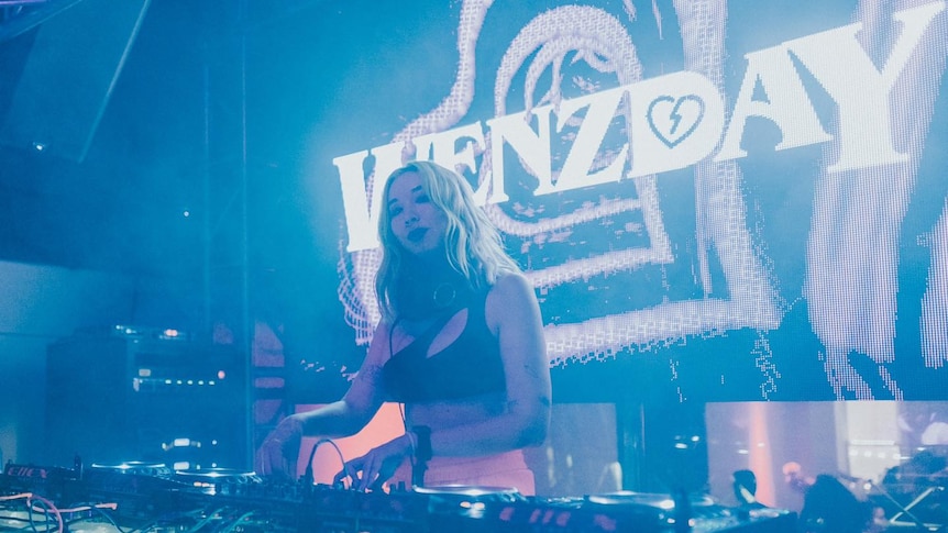 Wenzday has blonde hair and is DJing in a club. Her name is displayed on the screen behind her.