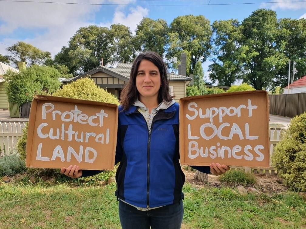A woman holds a cardboard sign in each hand.  One says 'protect cultural land' and the other says 'support local business'