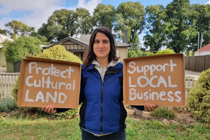 Una mujer sostiene un cartel de cartón en cada mano.  Uno dice 'proteger la tierra cultural' y el otro dice 'apoyar los negocios locales'