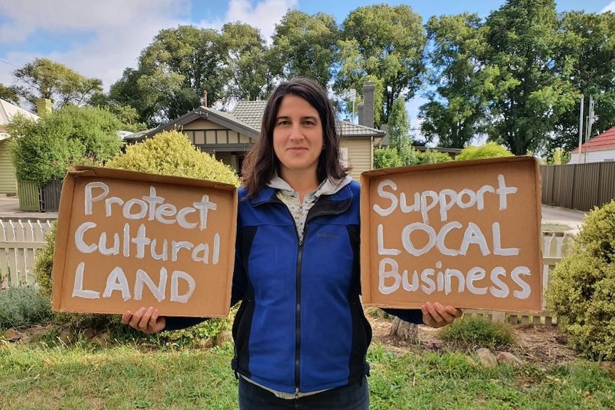 A woman holds a cardboard sign in each hand.  One says 'protect cultural land' and the other says 'support local business'