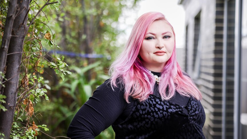 A woman with pink hair wearing a black dress stands in a garden with a faint smile as she looks to the camera