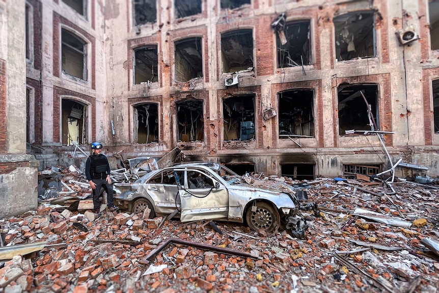a man walks by a destroyed car in a courtyard. He is surrounded by rubble and the ruins of a multi-storey building