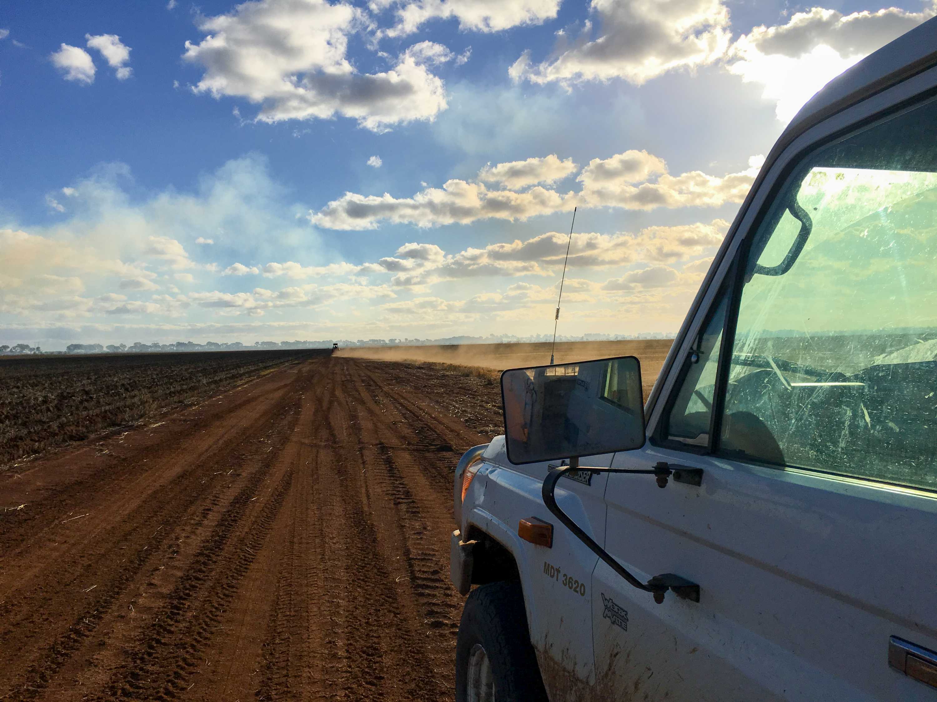 The passenger side of a ute in foreground with dust kicking up behind a vehicle on dirt road in the distance
