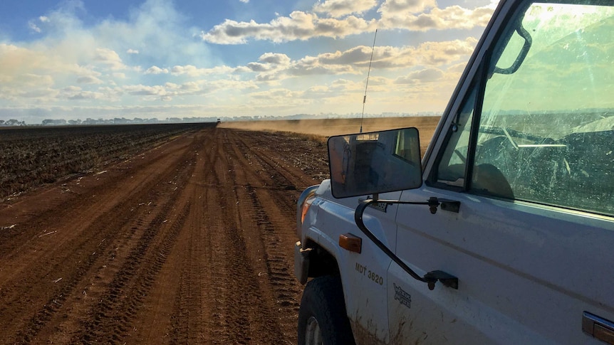 The passenger side of a ute in foreground with dust kicking up behind a vehicle on dirt road in the distance.