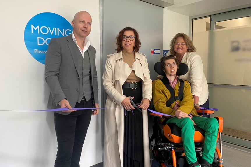 A man in a wheelchair poses with two women and another man while cutting a ribbon