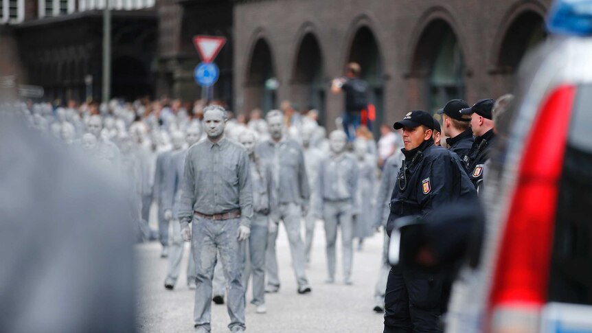 A crowd of people dressed and painted in grey walk by surveilling police