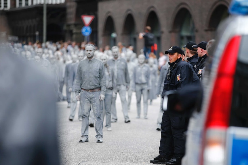 A crowd of people dressed and painted in grey walk by surveilling police