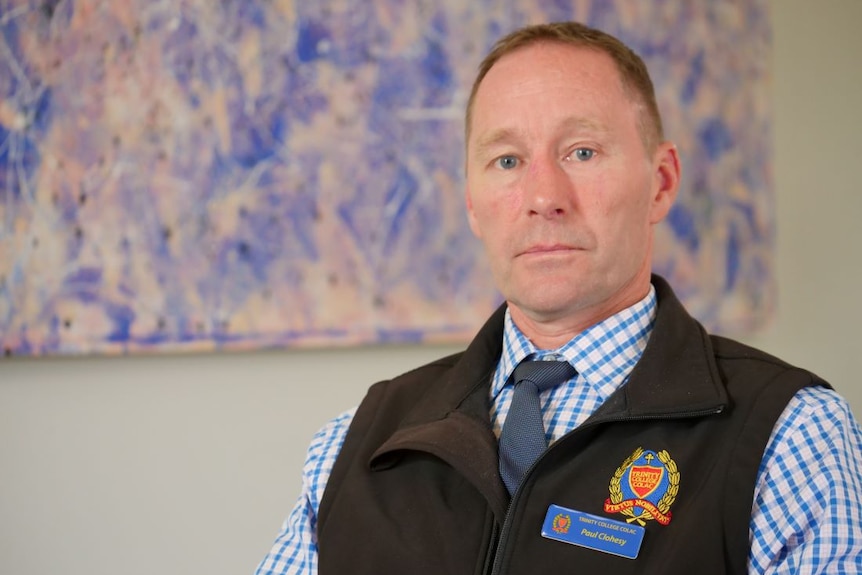 A man in a blue shirt, black tie and black vest with the Trinity College logo sits in front of a purple painting.