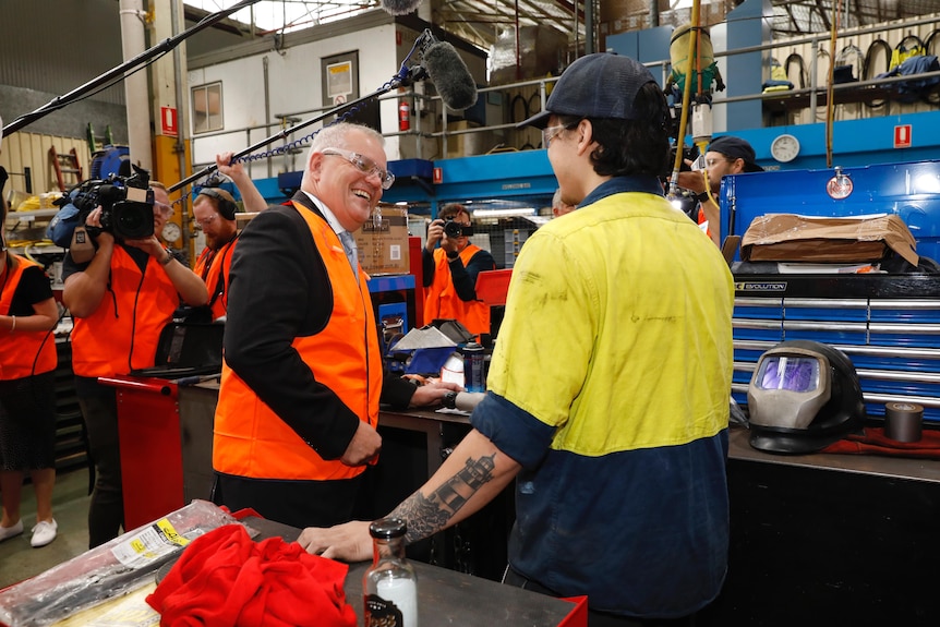 Scott Morrison wearing an orange hi-vis vest over his suit speaks to a worker wearing work gear and a cap.