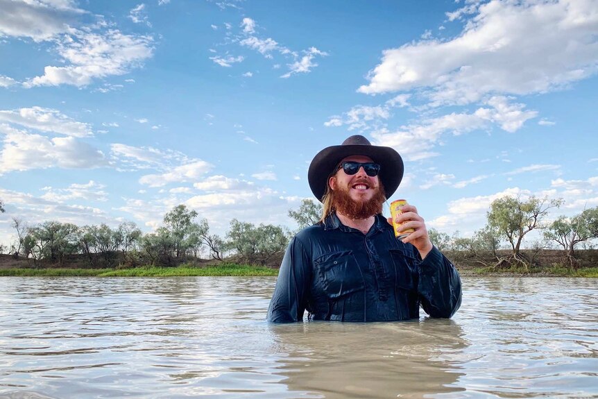 A smiling man stands holding a beer in a can in a river in the bush with water above his waist.