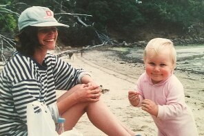 Photograph of a baby and woman on a beach smiling. There is a wooded headland behind them at the end of the beach.