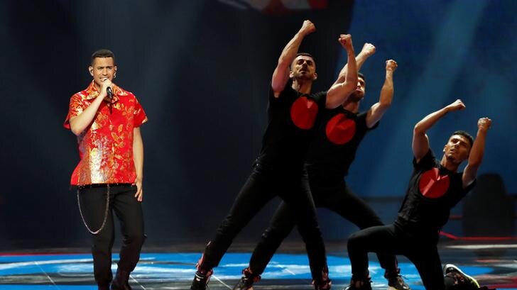 Mahmood, left, wears a red shirt as three dancers in black stand to his right. Mahmood is singing.