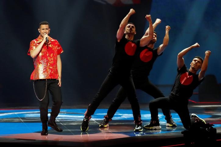 Mahmood, left, wears a red shirt as three dancers in black stand to his right. Mahmood is singing.