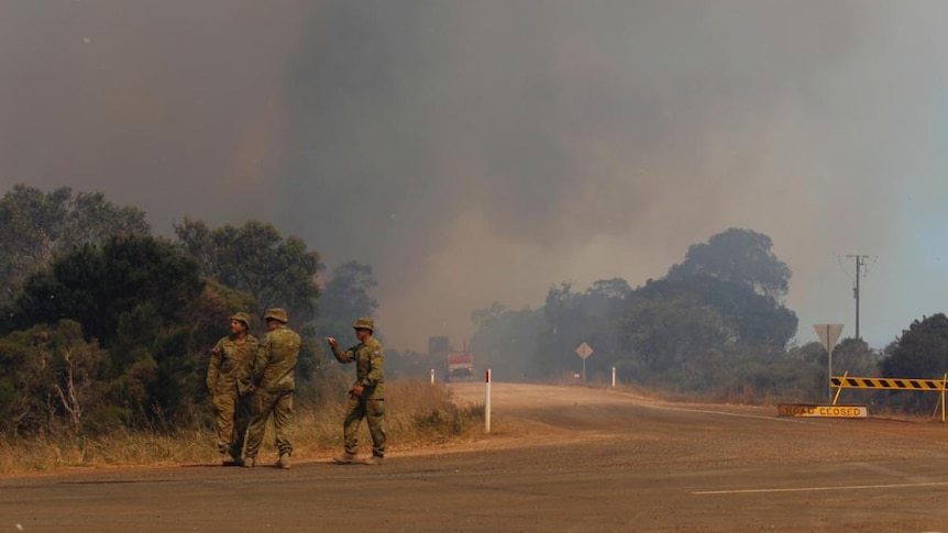 Three men in Army uniforms stand at a road block while the sky fills with thick black smoke from fire