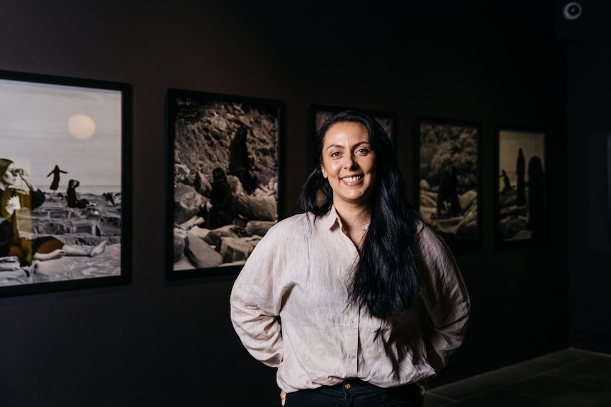 Hayley Millar Baker stands smiling in front of her Ramsay Art Prize entry, a set of photographs of women dressed in black