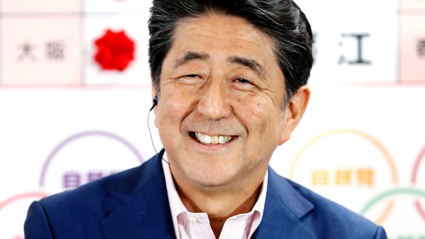 Shinzo Abe smiles in a navy blue suit with no tie in front of a whiteboard showing winning election candidates.
