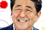 Shinzo Abe smiles in a navy blue suit with no tie in front of a whiteboard showing winning election candidates.