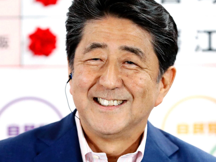 Shinzo Abe smiles in a navy blue suit with no tie in front of a whiteboard showing winning election candidates.