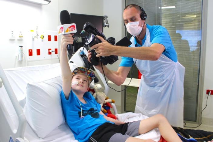 A boy in a hospital bed holds up a microphone as a man wearing a hospital mask and apron films him with a TV camera.