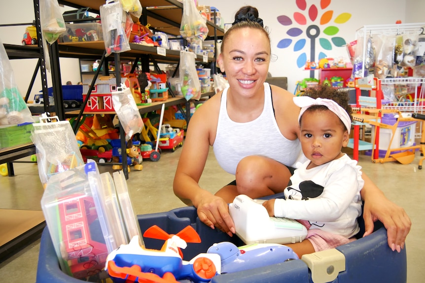 A smiling woman crouches behind a toddler in a cart filled with toys. There are shelves of toys in the background.