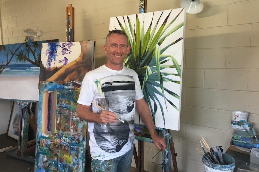 A man with paintings in the background. He is holding a paintbrush