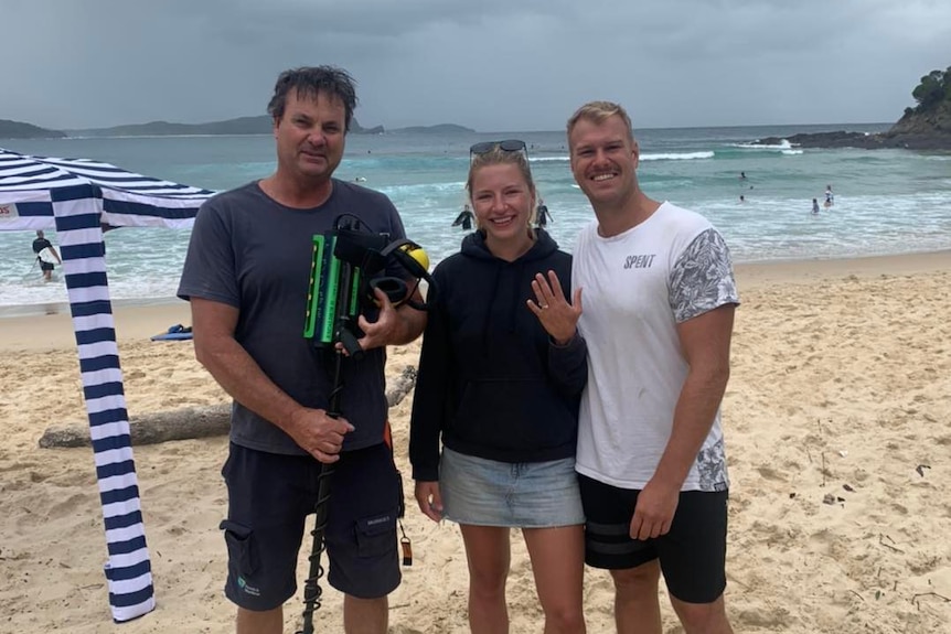 Un jeune couple se tient sur une plage, montrant une bague de fiançailles, à côté d'un homme avec un détecteur de métaux.