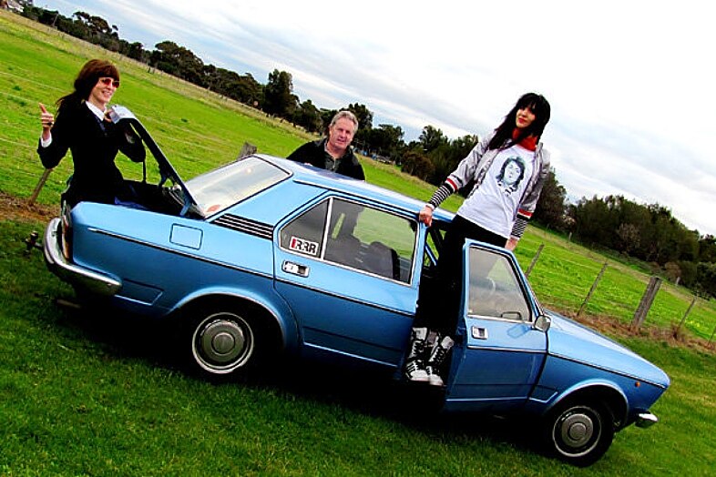 Three people pose with blue car, one in boot, one behind and one standing on the base of the driver door opening.