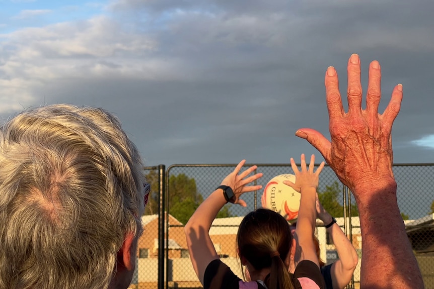 A woman with grey hair in the foreground with her hand up, and in the background is women playing netball.