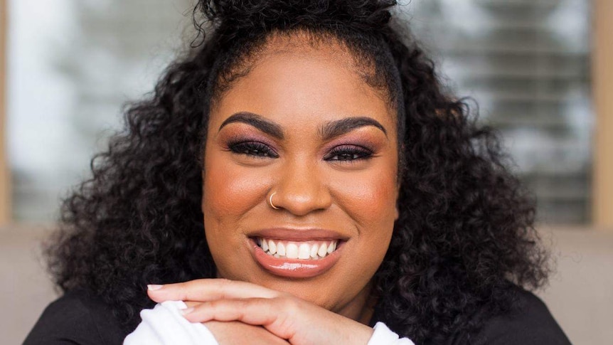 Young black woman writer Angie Thomas smiles directly at camera, hands at her chin, hair up on top of her head