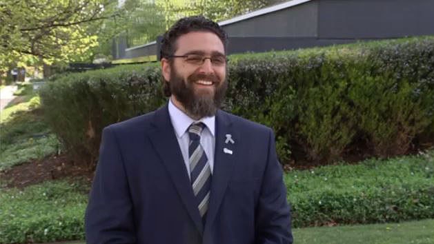 A man with a beard in a dark grey suit stands outdoors for an interview.