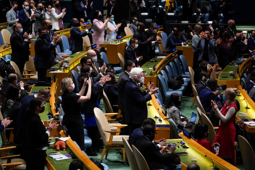 Delegates stand to applaud in the UN General Assembly chamber. Some are filming on their smartphones. 