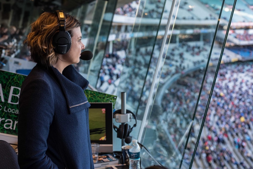 Kelli Underwood wearing headset standing in ABC commentary box with MCG crowd in background.