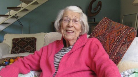 An elderly lady sits on a couch in a living room and smiles.