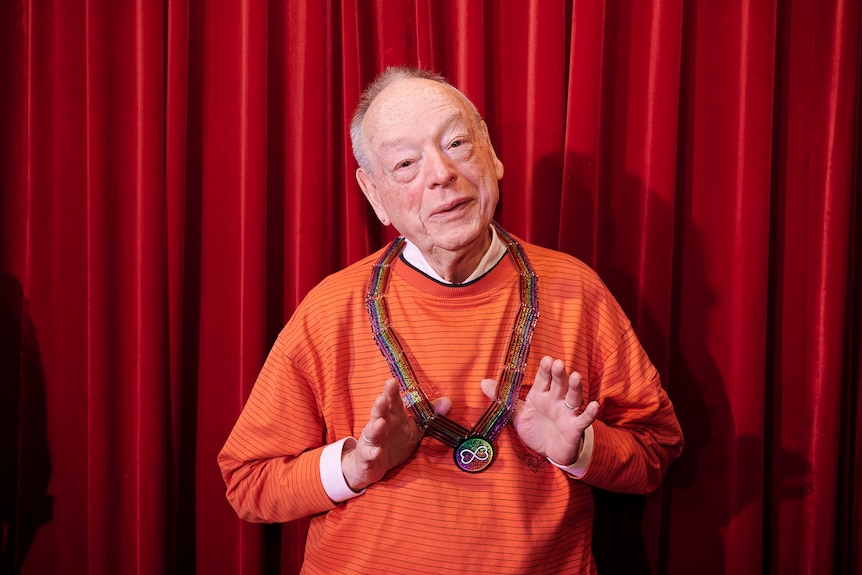 An older man wearing an striped orange jumper has his thumbs holding out the very colourful WorldPride medallion he is wearing. 
