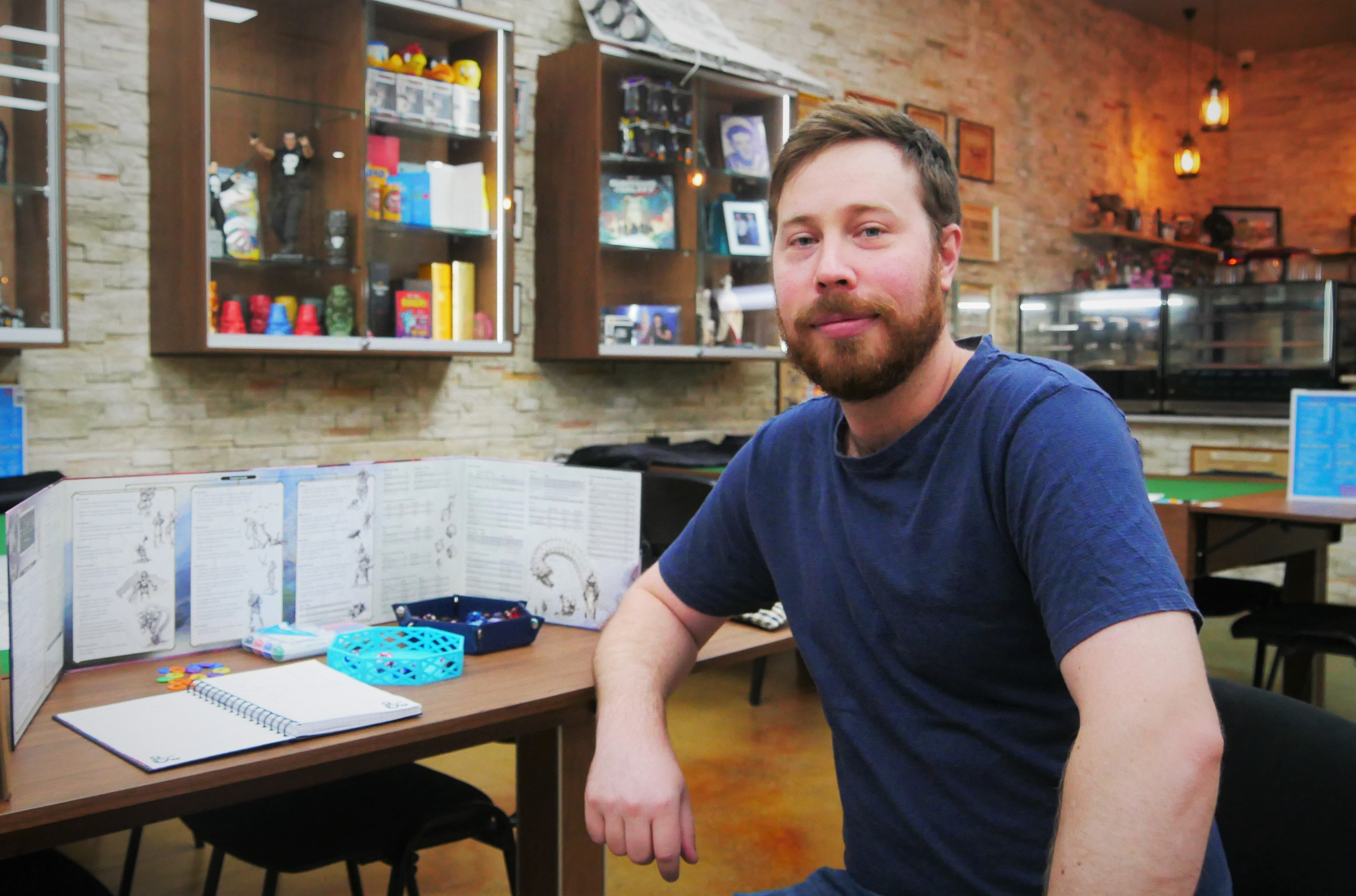 A man sits at a table, he looks happily at the camera, on the table behind him books and equipment to play a game