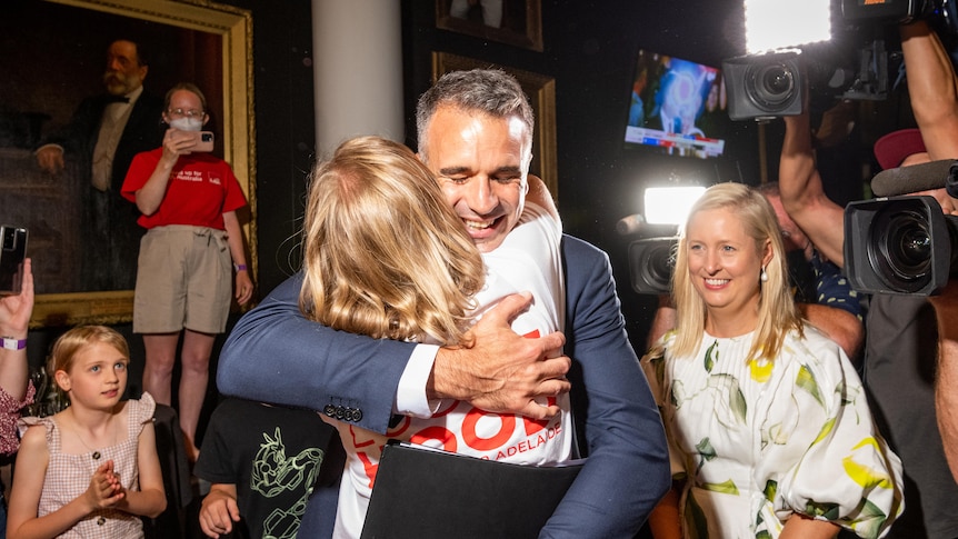 A smiling man hugs a woman, surrounded by people and cameras