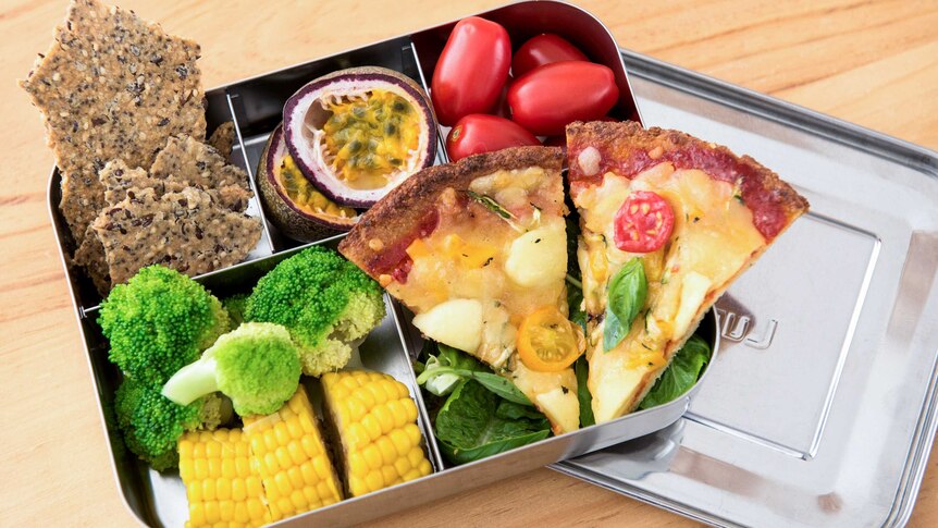 Homemade pizza in a bento box with tomatoes, corn, broccoli, homemade crackers and passionfruit.