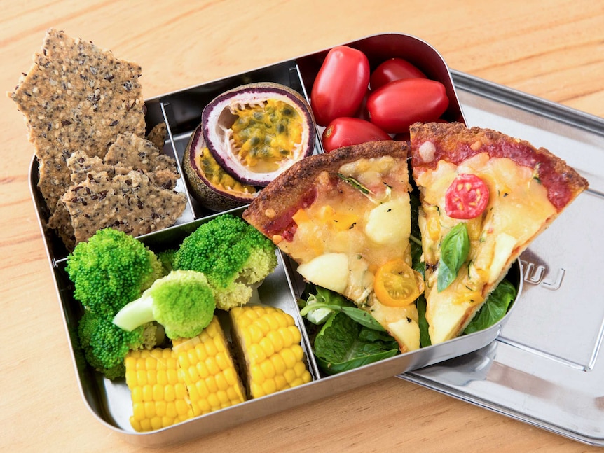 Homemade pizza in a bento box with tomatoes, corn, broccoli, homemade crackers and passionfruit.