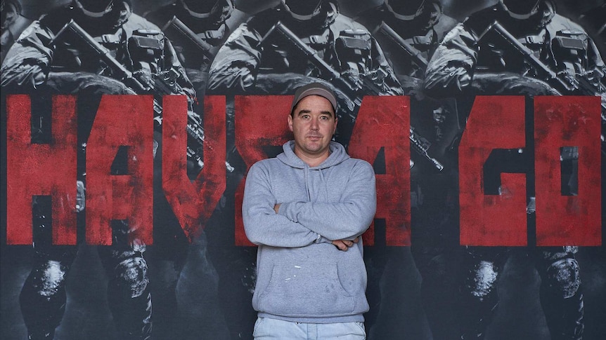A man with a grey hoodie standing in front of a large piece of art featuring officers with their guns behind the words have a go