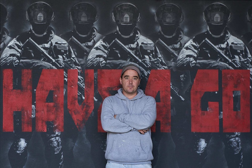 A man with a grey hoodie standing in front of a large piece of art featuring officers with their guns behind the words have a go