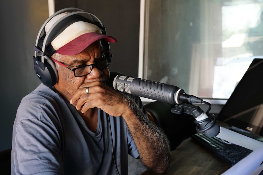 Warren H Williams wearing headphones in front of a microphone, looking down at a piece of paper with hand over his mouth