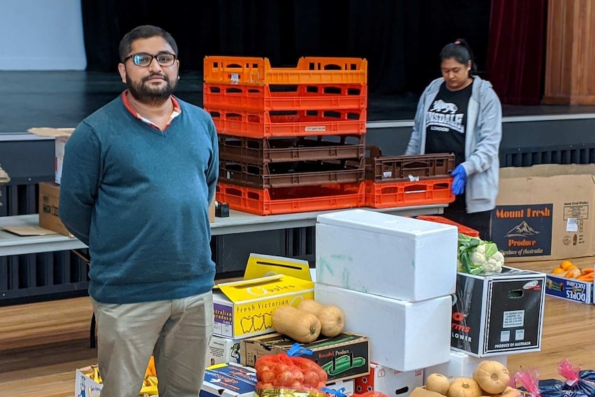 A man wearing a blue jumper stands in a hall with his hands behind his back with boxes of food on the floor next to him.