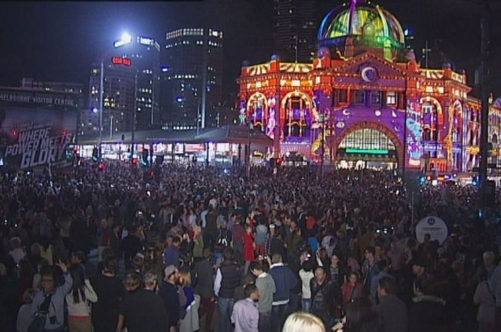 Huge crowds of people fill the intersection in front of Flinders Street Station, the exterior of which is illuminated in light.