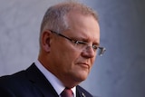 Man with grey hair and glasses looking down with a blank or sad expression.