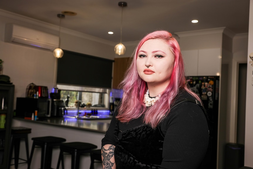 A woman with pink hair looking sad sitting in a kitchen.