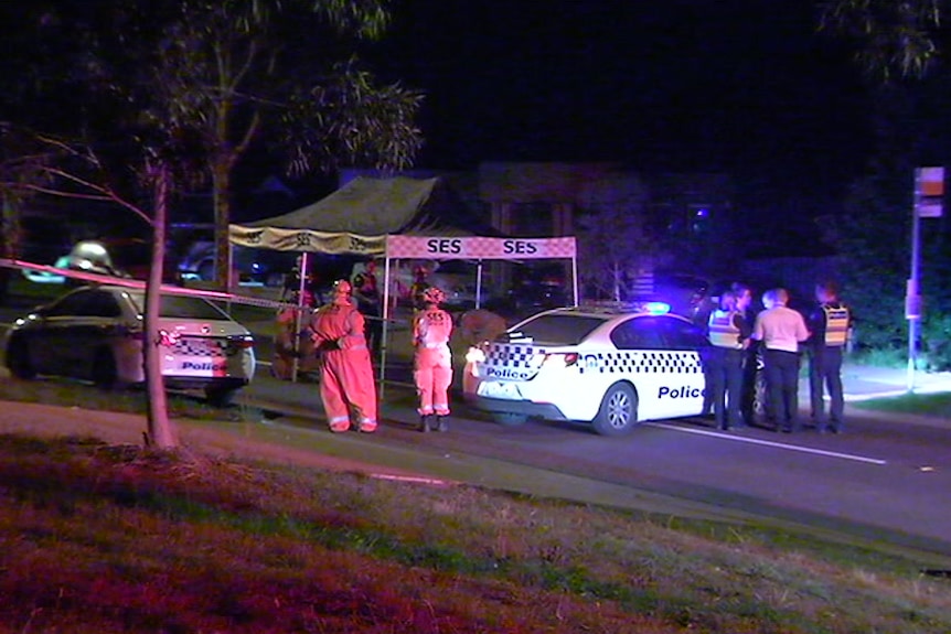 Several police and SES personnel stand near two police cars and an SES tent at night in a suburban street.