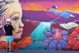 An artist works on a mural which shows a young girl looking out at flowers and books.